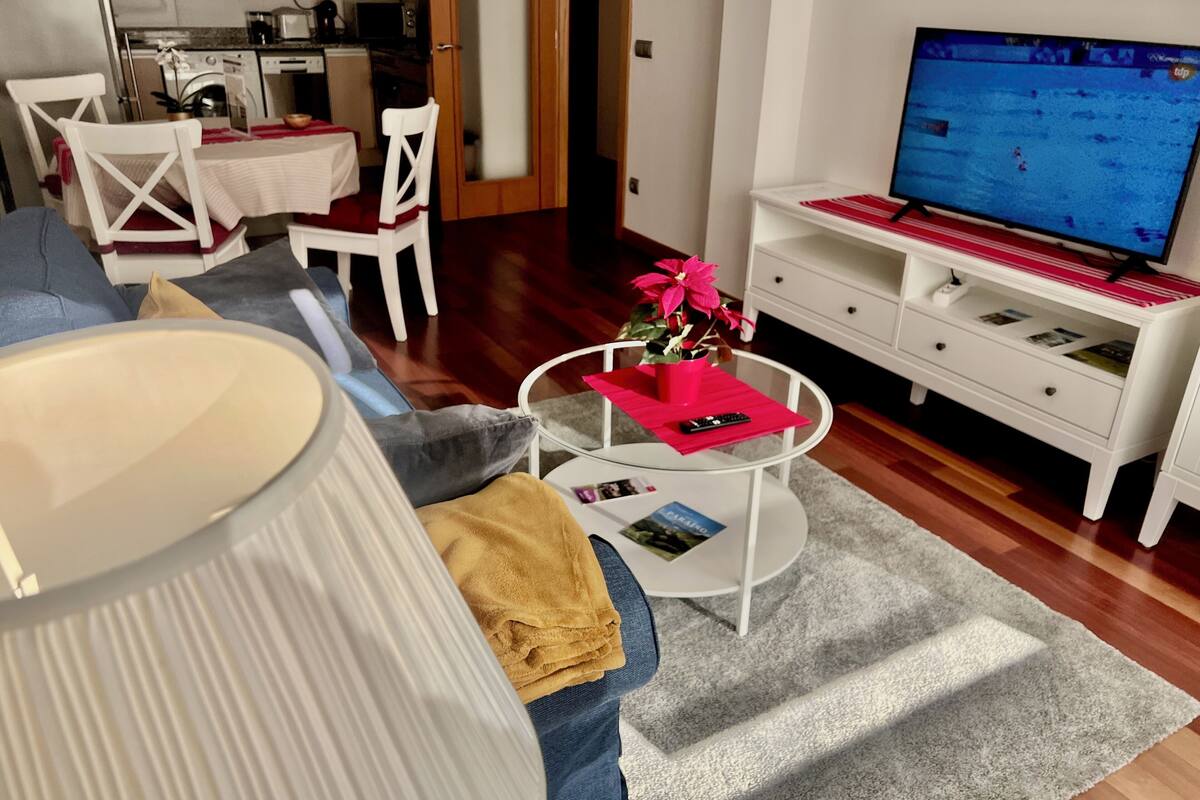 Un salon moderne avec un canapé, une table basse ornée d’un vase de fleurs, un téléviseur et, au fond, un coin cuisine.