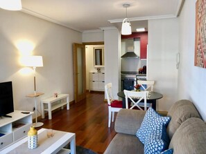 A living room with a sofa, a TV, a lamp, and a dining table with chairs. There is a kitchen in the background with a stove and cabinets. - Apartamento Europa Prados - Atenea (Oviedo)