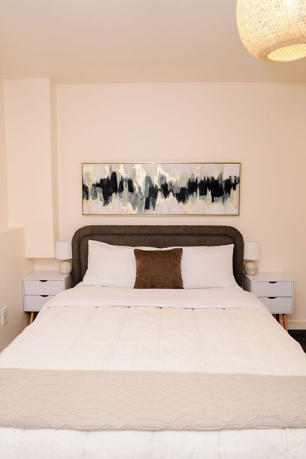 A modern bedroom with a large bed, a headboard with a textured design, a wall-mounted abstract painting, and two bedside tables with lamps.