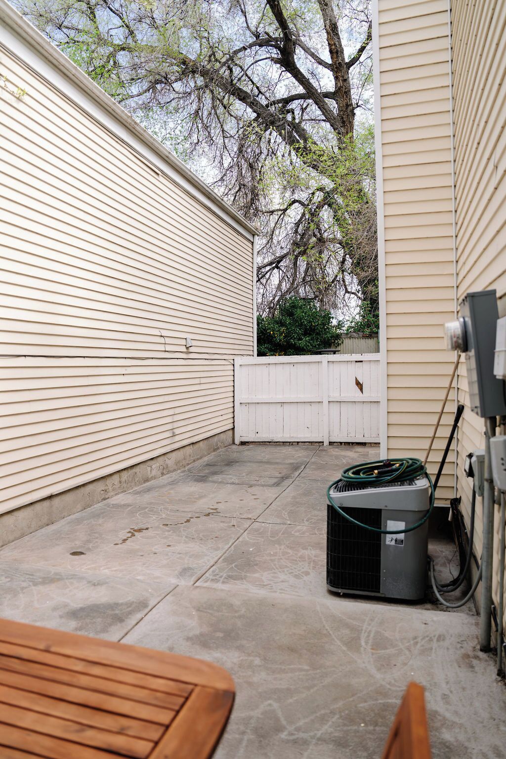 A covered patio area with a concrete floor, a wooden bench, a white fence, and a large tree in the background.
