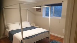 Een hemelbed met wit beddengoed, een klein nachtkastje en een raam met uitzicht op bomen.
