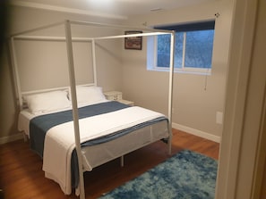 A four-poster bed with white bedding, a small bedside table, and a window with a view of trees. - Beautiful 2-bedroom basment house with your own entrance (Sandy)