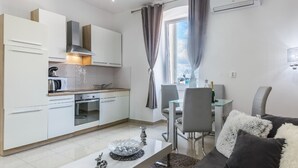 A modern kitchen with white cabinets, a built-in oven, and a coffee maker. There is a dining area with a table and chairs, and a sofa with fluffy pillows. - Comfrot One Bedroom Apartment with Sea View (Pula)