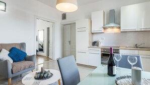 Un salon moderne avec un canapé, une table basse et une table à manger. La pièce comprend une cuisine avec des meubles blancs et une cuisinière. Une grande fenêtre laisse entrer la lumière naturelle.