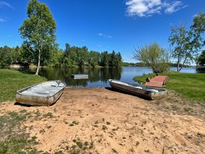Un paisaje junto al lago con dos barcos amarrados en una orilla arenosa, rodeado de árboles y un cielo despejado.