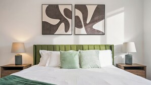 Un lit bien fait, avec une tête de lit verte, deux tables de chevet avec des lampes et des œuvres d’art abstraites accrochées au mur.