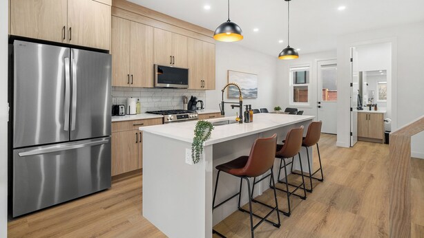 Une cuisine moderne dotée d’appareils électroménagers en acier inoxydable, de meubles de rangement en bois, d’un îlot central avec des tabourets de bar, d’un éclairage suspendu et d’une vue sur la salle à manger adjacente.