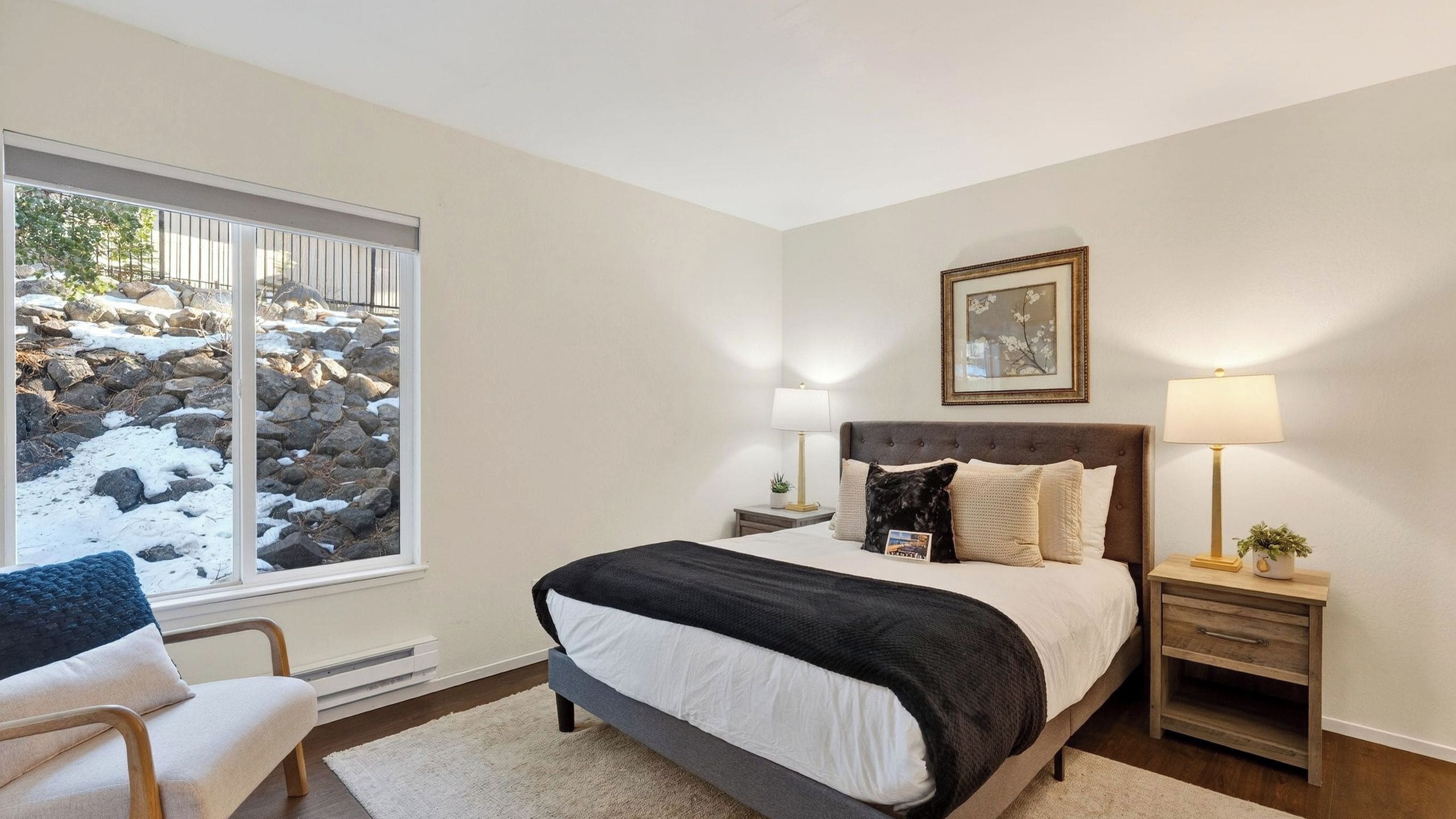 A bedroom with a large window, a bed, a bedside table with a lamp, and a chair.
