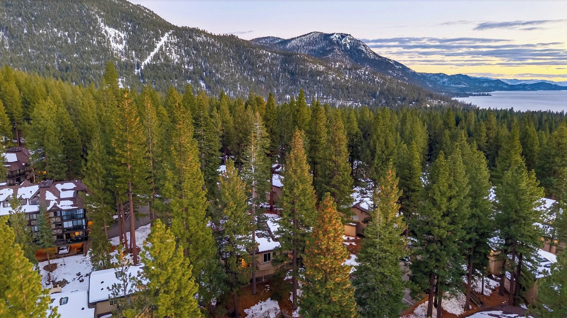 A lodge surrounded by dense evergreen forest, with snow on the ground and mountains in the background.