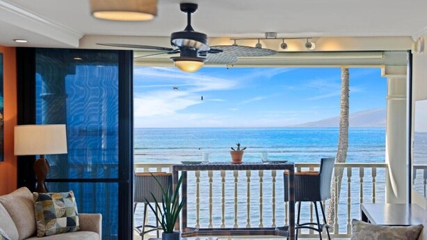 A room with a balcony overlooking the ocean, a ceiling fan, a table with chairs, and a potted plant.
