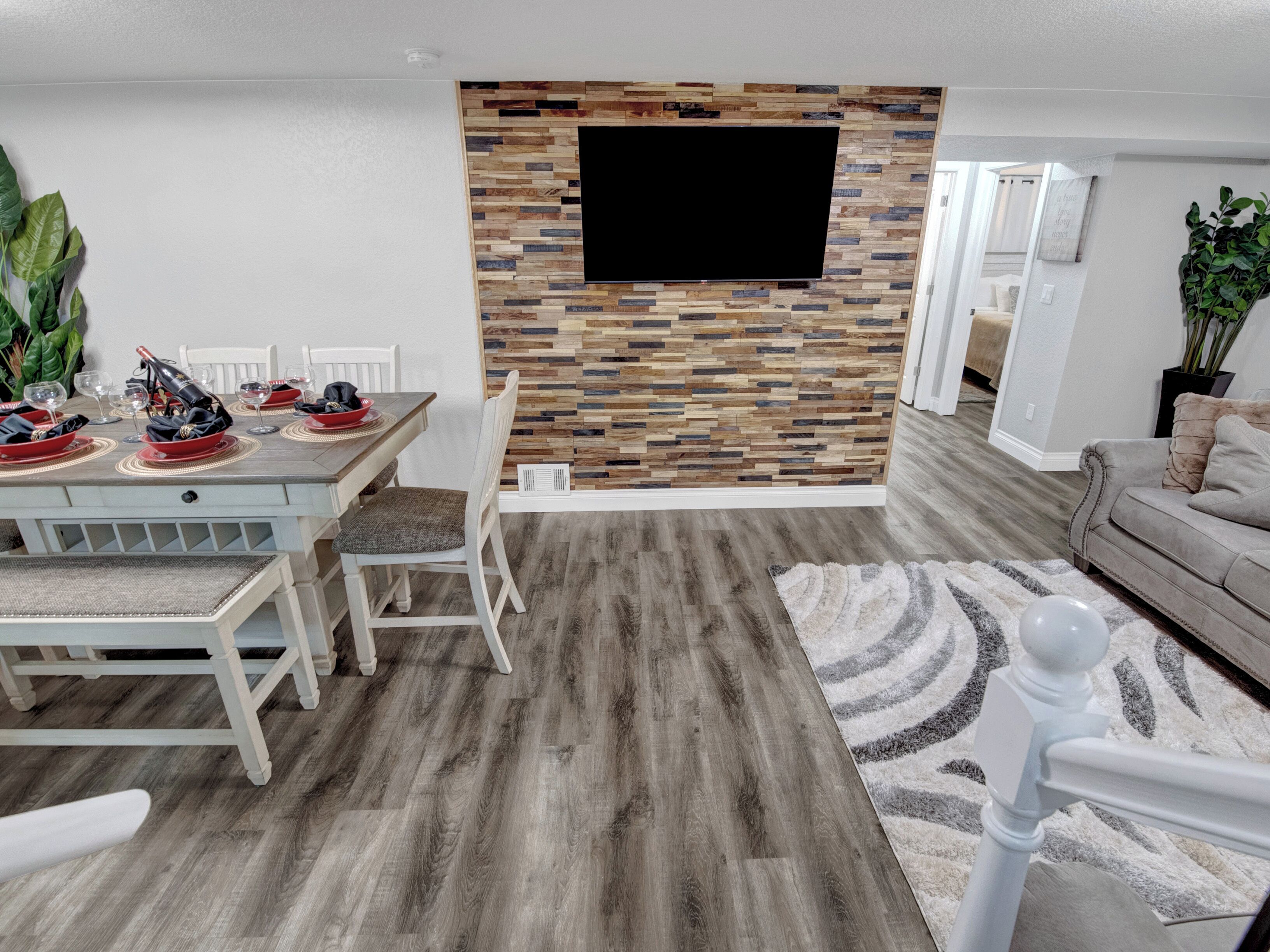 A dining area with a wooden table set for four, a flat-screen TV mounted on a wooden wall, and a grey carpet leading to a sofa.