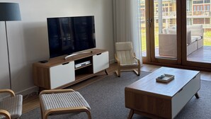 A modern living room with a flat-screen TV, a sofa, a coffee table, and a chair. There is a large window with a view of a building and a balcony.