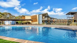 A swimming pool with a clear blue water surface, surrounded by a paved deck. In the background, there are buildings with a modern architectural style, featuring a mix of flat and sloped roofs.