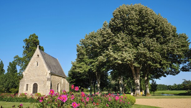 Un edificio de piedra con techo triangular, rodeado de un jardín bien cuidado con flores rosadas y árboles grandes.