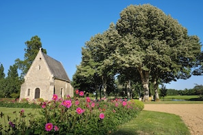 Un edificio de piedra con techo triangular, rodeado de un jardín bien cuidado con flores rosadas y árboles grandes.