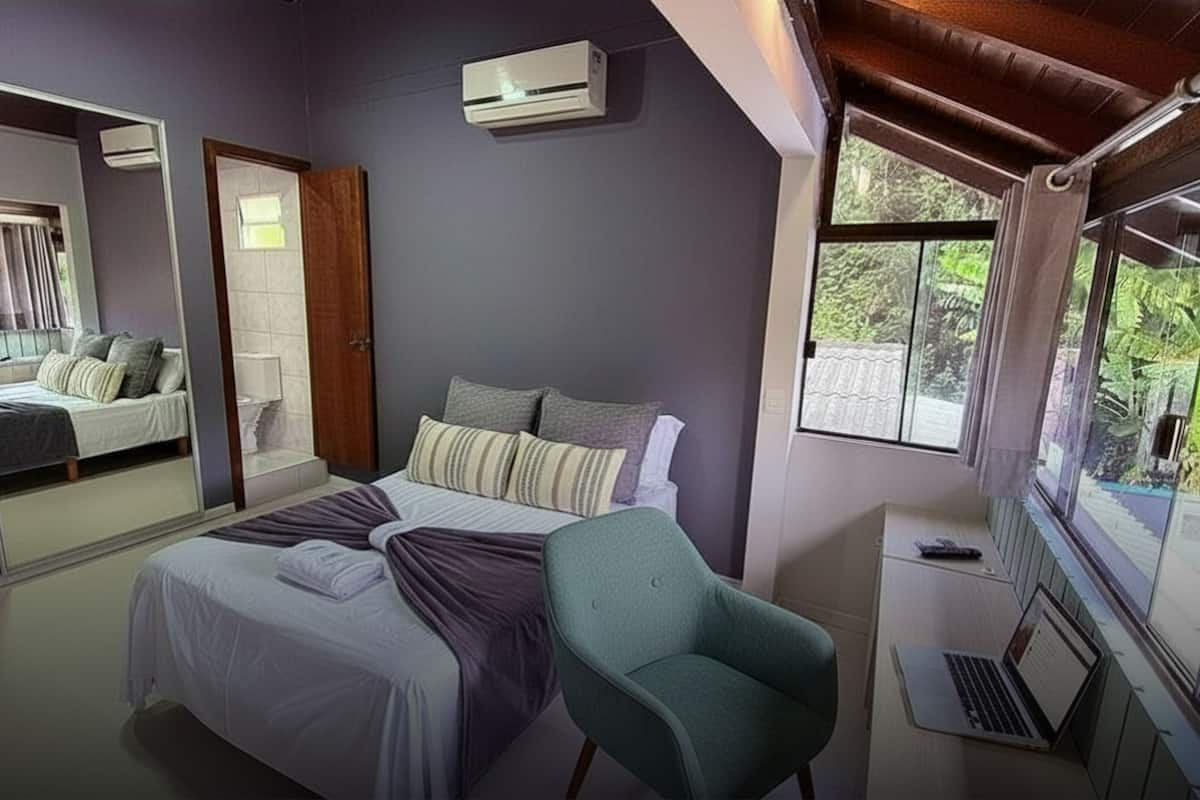 A bedroom with a bed, a chair, a desk with a laptop, a mirror, and a window with a view of greenery.