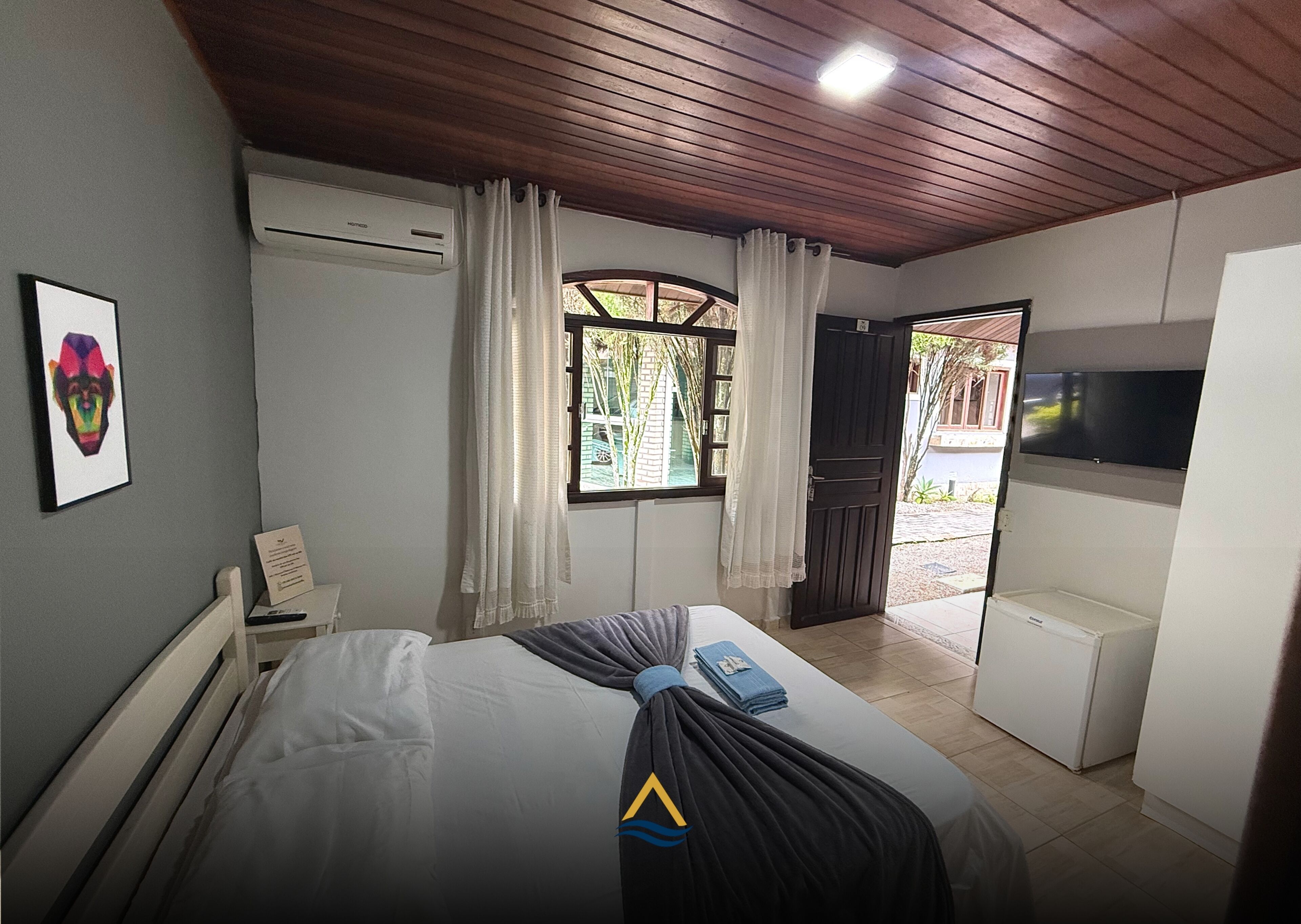 A bedroom with a bed, a TV, an air conditioner, a window with curtains, and a door leading outside.