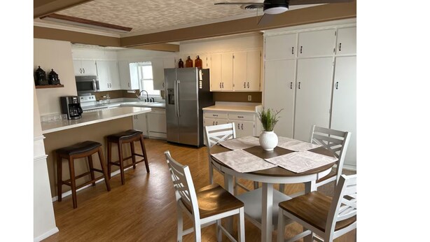 A modern kitchen with white cabinets, a stainless steel refrigerator, wooden stools, and a dining table with chairs. - My family’s little nest, where our kids grew. (Perryton)