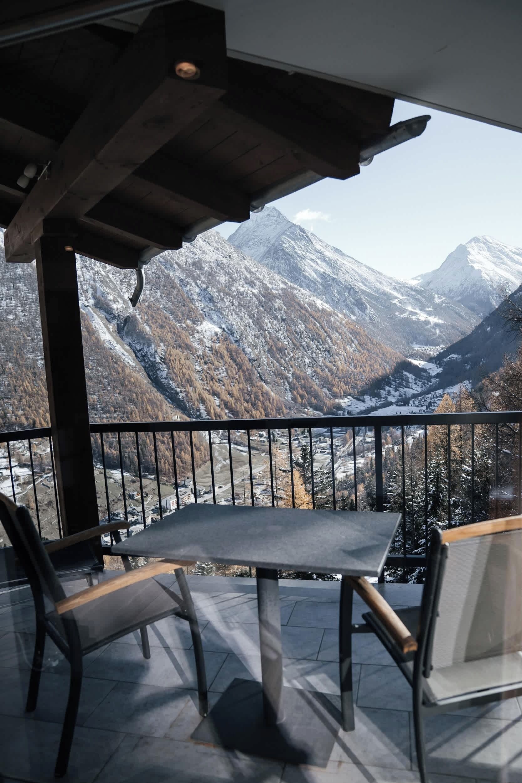 A balcony with a table and chairs, overlooking a mountainous landscape with snow-covered peaks and a valley below.
