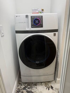 Een compacte, moderne wasmachine met voorlader en een digitaal bedieningspaneel, geplaatst in een kleine wasruimte met betegelde vloer.