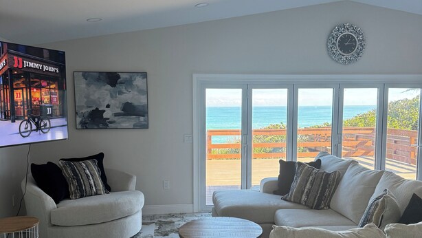 Een woonkamer met een groot raam met uitzicht op een strand, een wandtelevisie en een ronde salontafel.