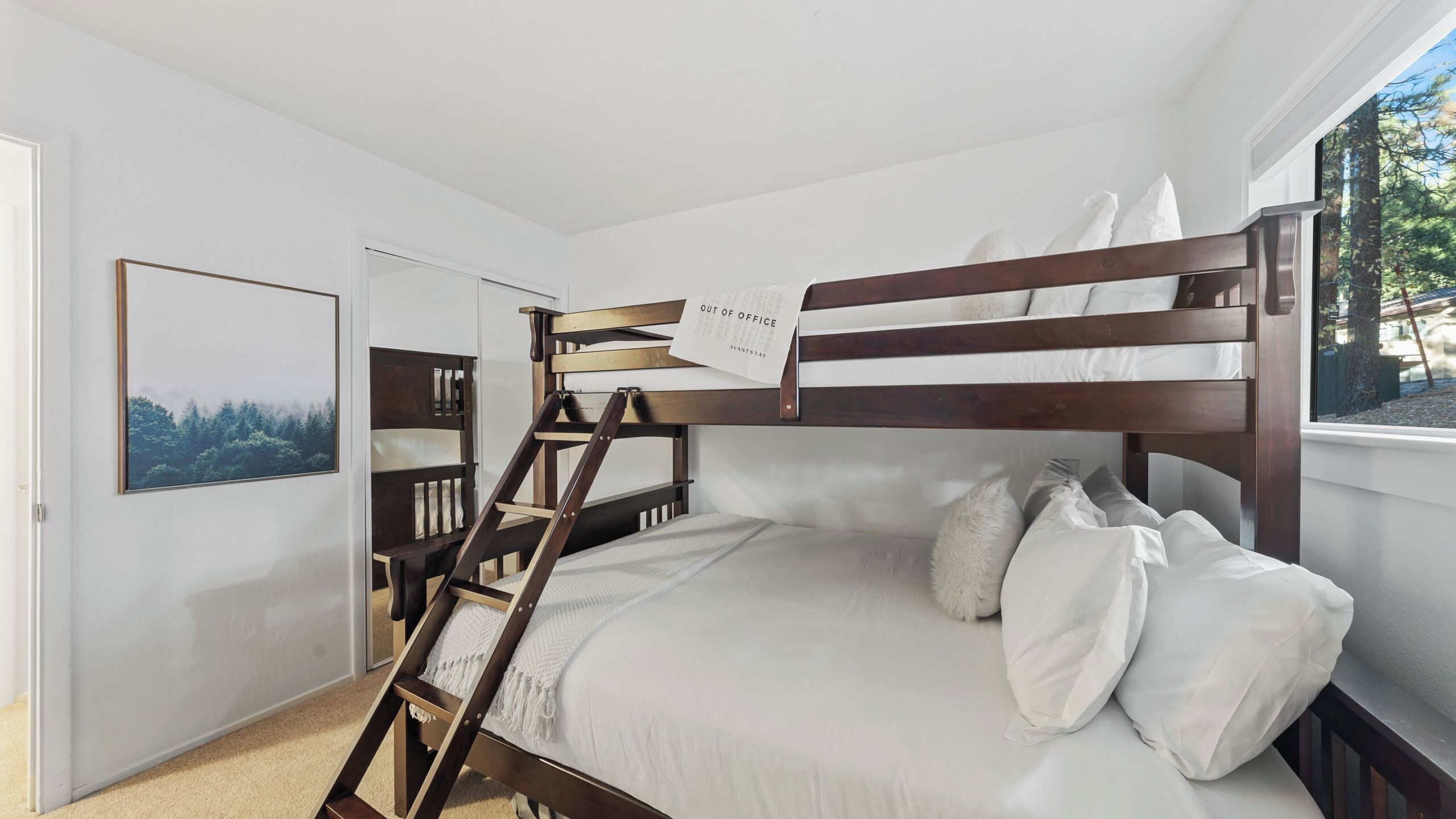 A bunk bed room with a ladder, a framed picture on the wall, and a window with a view of trees.