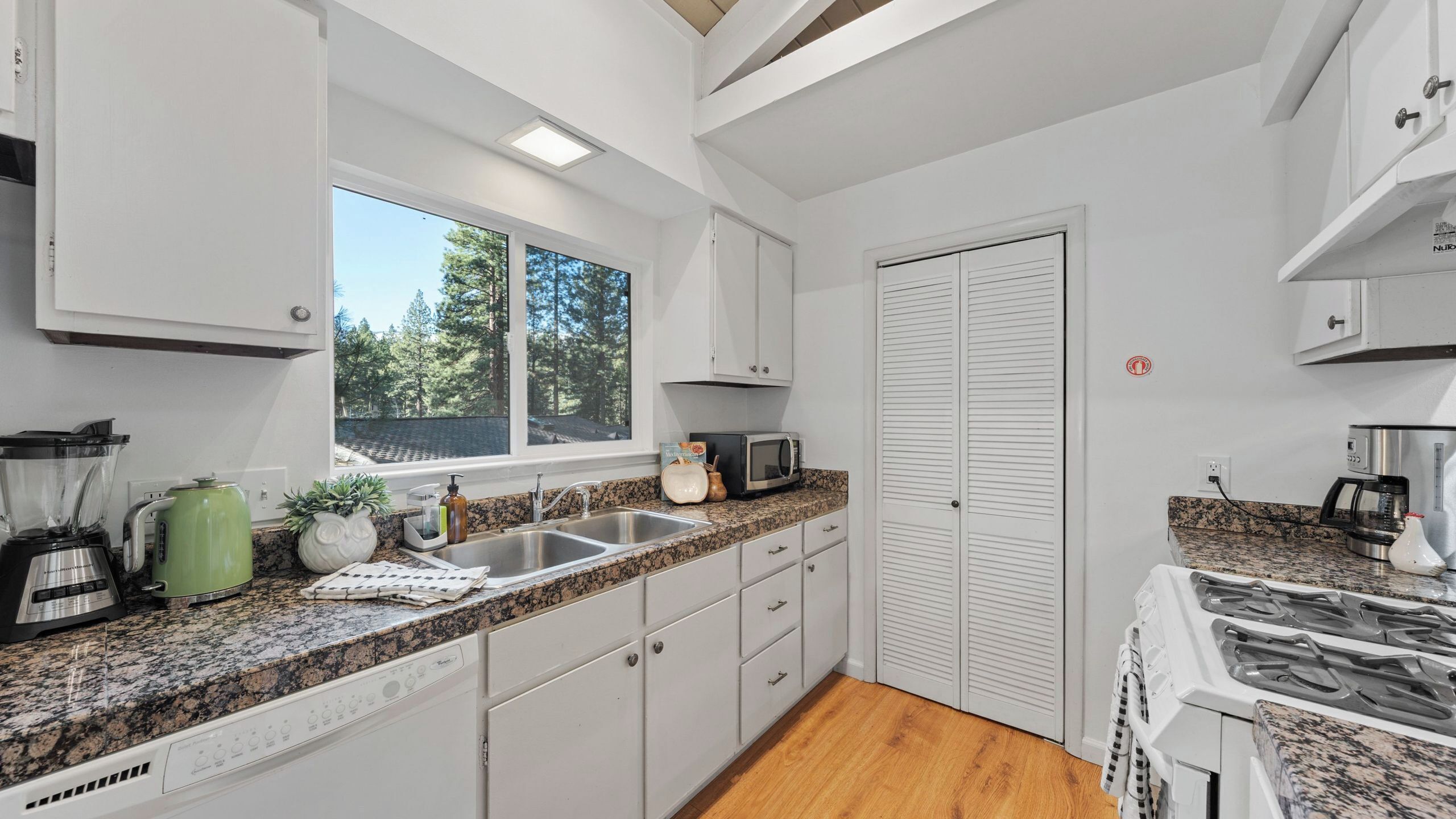 A modern kitchen with white cabinets, granite countertops, a built-in stove, and a window offering a view of trees.