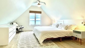 Ein Schlafzimmer mit schrägem Dach, einem Bett mit weißer Bettwäsche, einem Nachttisch, einem Fenster mit Aussicht und einem Deckenventilator.