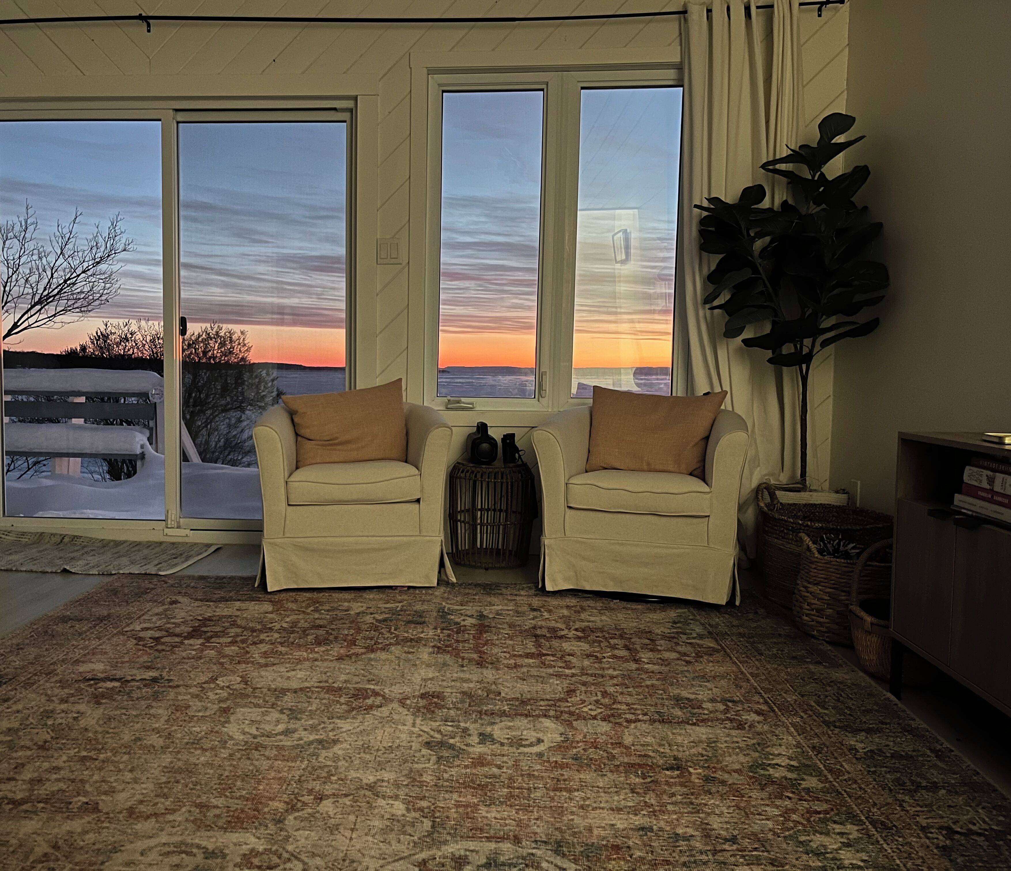 Un salon avec un canapé beige, une grande plante en pot et une vue sur un coucher de soleil à travers de grandes fenêtres.