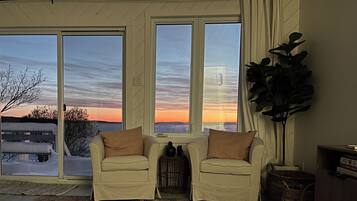 Un salon avec un canapé beige, une grande plante en pot et une vue sur un coucher de soleil à travers de grandes fenêtres.