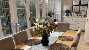 Een eetgedeelte met een lange houten tafel, beiges bekleden stoelen, een vaas met bloemen en uitzicht op groen door grote ramen.