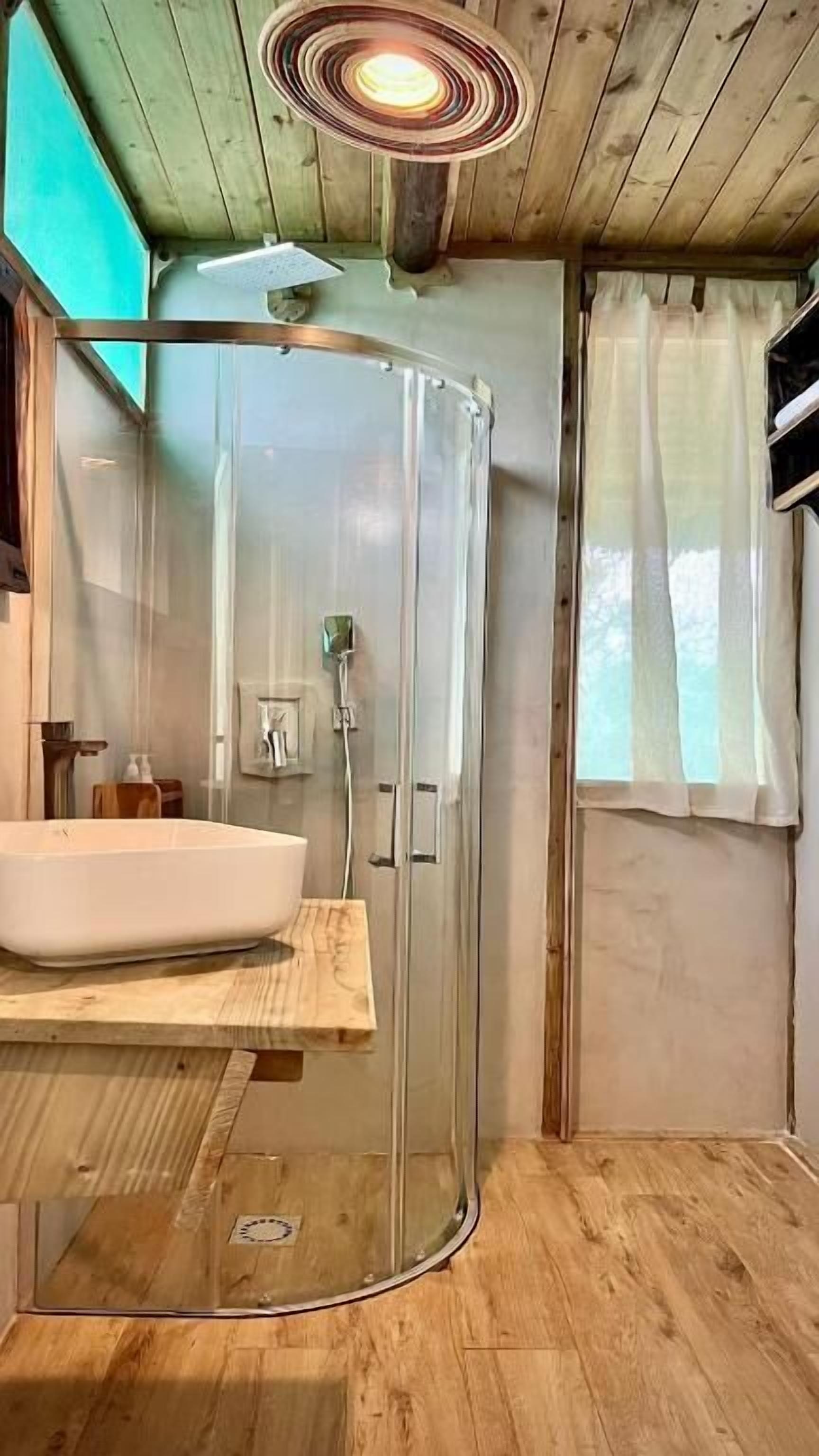 A modern bathroom with a glass-enclosed shower, a wooden vanity with a sink, and a window with sheer curtains.