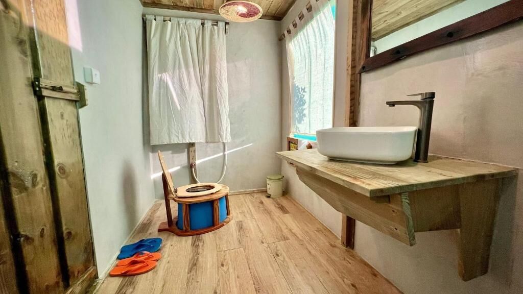 A small bathroom with a wooden floor, a white sink, a wooden chair, and a window with curtains.