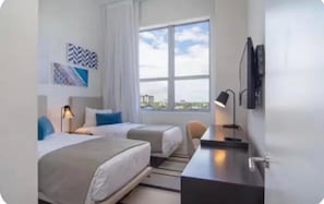 Ein Hotelzimmer mit zwei Betten, einem Schreibtisch, einem Stuhl, einem Fernseher und einem Fenster mit Blick auf die Stadt.