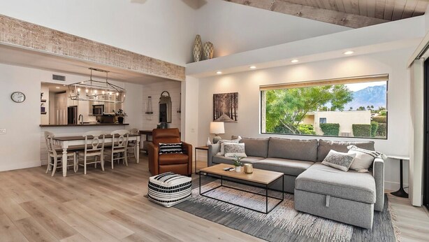 A spacious living area with a sofa, coffee table, and dining table. A kitchen is visible in the background, and a large window offers a view of greenery and mountains. - STUNNING FURNISHED POOLSIDE HOME AT MISSION HILLS COUNTRY CLUB RANCHO MIRAGE (Rancho Mirage)