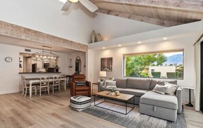 A spacious living area with a sofa, coffee table, and dining table. A kitchen is visible in the background, and a large window offers a view of greenery and mountains. - STUNNING FURNISHED POOLSIDE HOME AT MISSION HILLS COUNTRY CLUB RANCHO MIRAGE (Rancho Mirage)