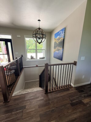 Una escalera con barandilla de madera, un candelabro, una ventana con vista a follaje y un cuadro en la pared.