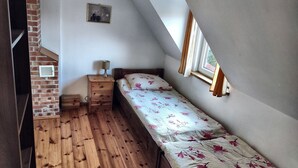 Ein kleiner, schräger Raum mit Holzboden, einem Einzelbett mit floralem Bettbezug, einem Nachttisch mit Lampe, einem kleinen Bilderrahmen und einem Fenster mit Vorhängen.