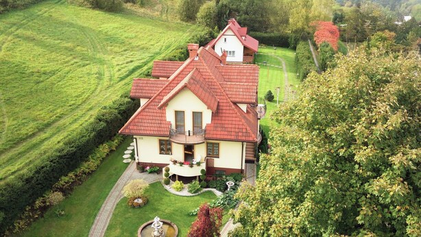 Ein zweistöckiges Haus mit rotem Ziegeldach, einem Balkon und einem gepflegten Garten mit Brunnen.