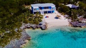 Eine Luftaufnahme einer Strandlage mit einem großen Haus, klarem türkisblauem Wasser und üppiger Vegetation.