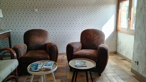 Ein Zimmer mit zwei braunen Ledersesseln, einem kleinen runden Tisch mit Zeitschriften, raumschmückendem, gemustertem Wandpapier und einem Fenster mit Vorhängen.