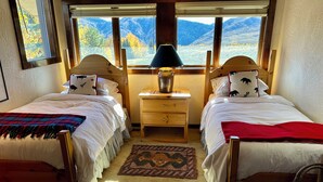 A room with two beds, a nightstand with a lamp, a rug, and a view of mountains through large windows. - Private 4 Bedroom 4 Bath Sun Valley Getaway. Hot Tub/Large Yard/ Dog Friendly (Sun Valley)