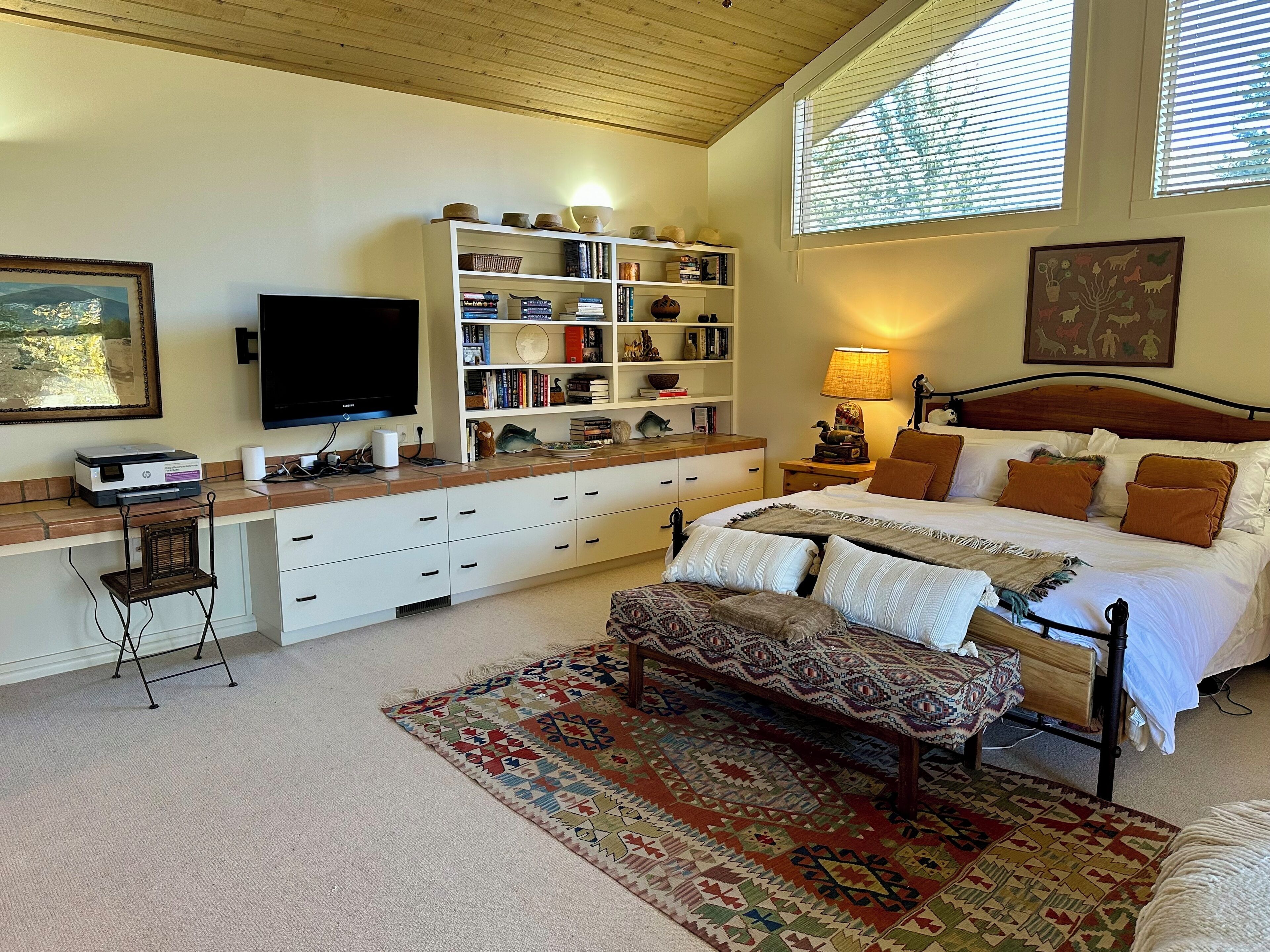 A bedroom with a bed, bedside tables, a desk with a computer, a bookshelf, and a window with blinds.