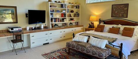 A bedroom with a bed, bedside tables, a desk with a computer, a bookshelf, and a window with blinds.