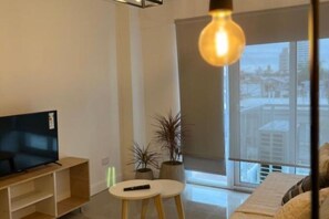 Un saló modern amb televisió de pantalla plana sobre un suport de fusta, una taula rodona blanca amb un comandament a distància i dues plantes en testos.