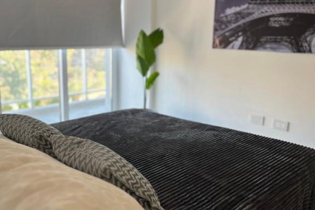 Un letto con un cuscino a fantasia e una coperta scura e strutturata in una stanza con una finestra, una pianta e un interruttore della luce fissato al muro.