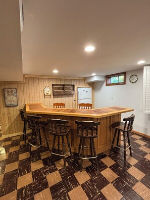 A bar area with a wooden counter and four stools, a refrigerator, and a clock on the wall. - Travel Team Retreat: Fabulous 4-bedroom house with AC in charming Dyersville (Dyersville)