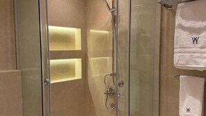 Ducha con cerramiento de vidrio, cabezal de ducha y panel de control cromados, paredes con azulejos color beige y dos toallas blancas con un monograma colgadas en un perchero.