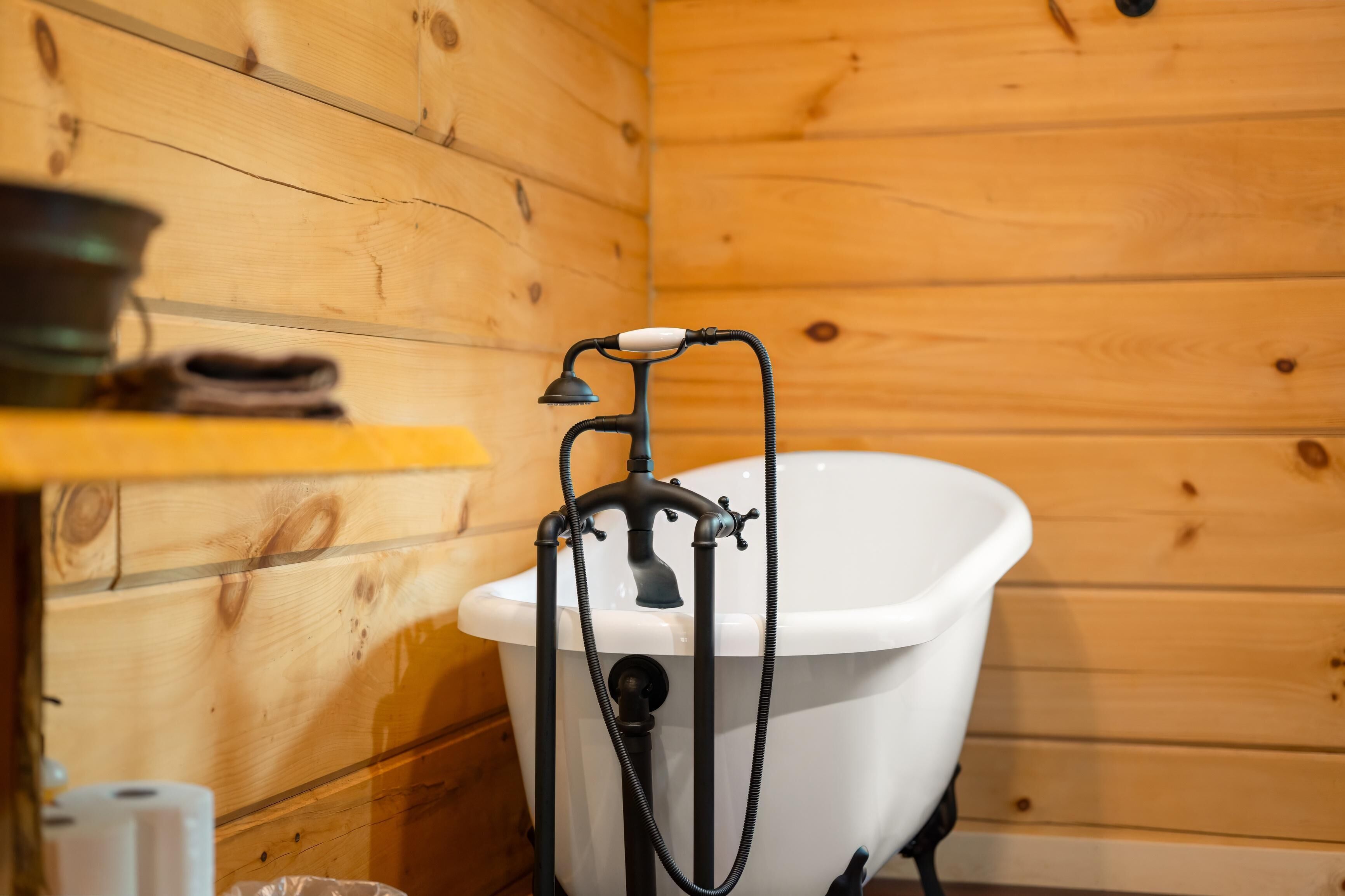 A wooden cabin bathroom with a white clawfoot bathtub, a wooden shelf with a metal pot, and a black hose attached to the tub.