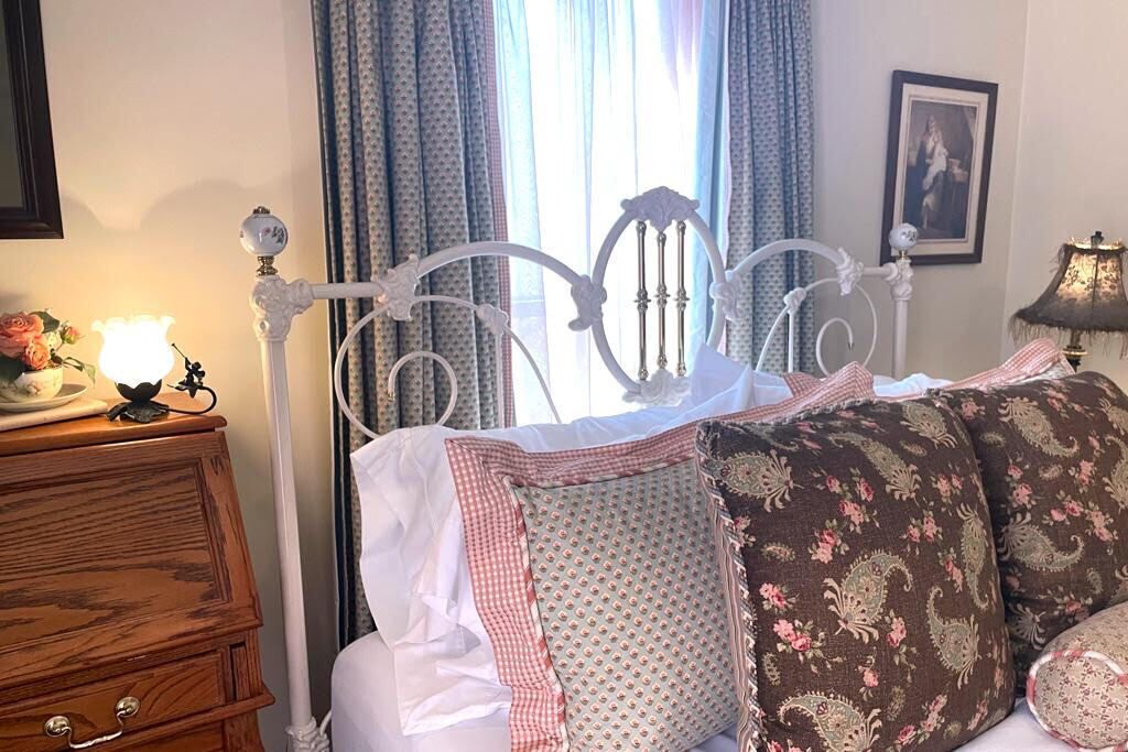 A bed with a white metal frame, a wooden dresser, a lamp, and a window with curtains.